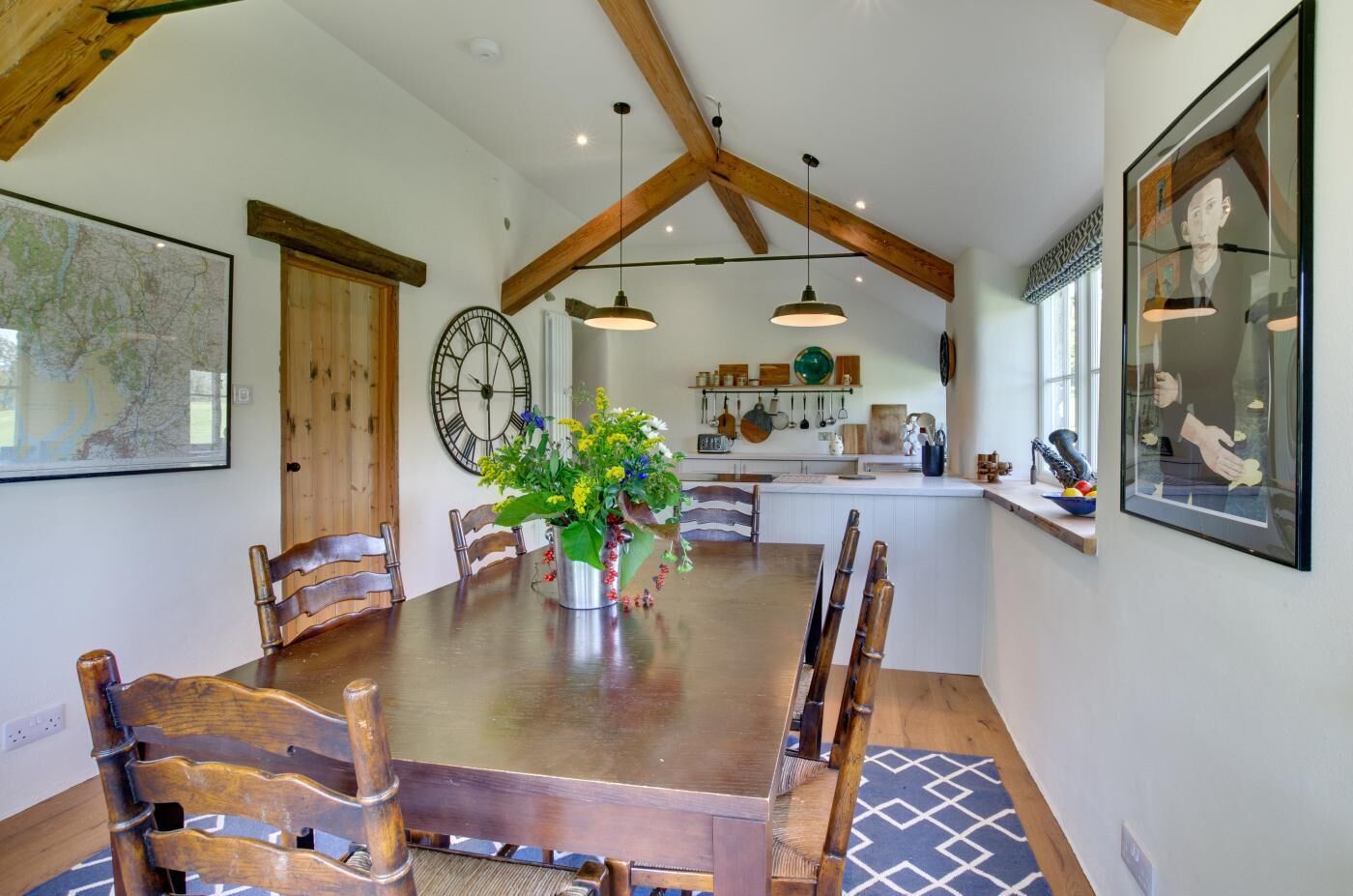 A dining area with a wooden table, chairs, a clock, and a vase of flowers.