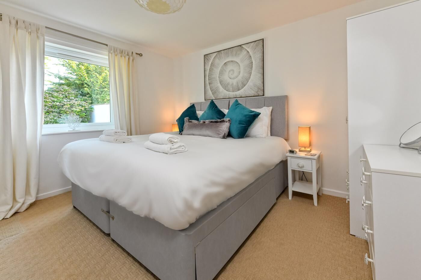 A neatly made bed with white linens and teal pillows, a bedside lamp, and a view of greenery outside.