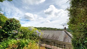 A stone house with a sloped roof, surrounded by lush greenery and flowering plants. - 2 Bed in Fowey (oc-secret) (Fowey)
