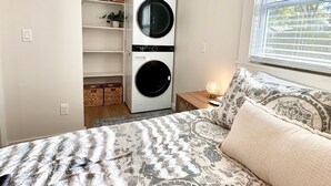 Ett sovrum med en säng, en väggmonterad tvättmaskin och torktumlare, ett fönster med persienner och en hylla med korgar.