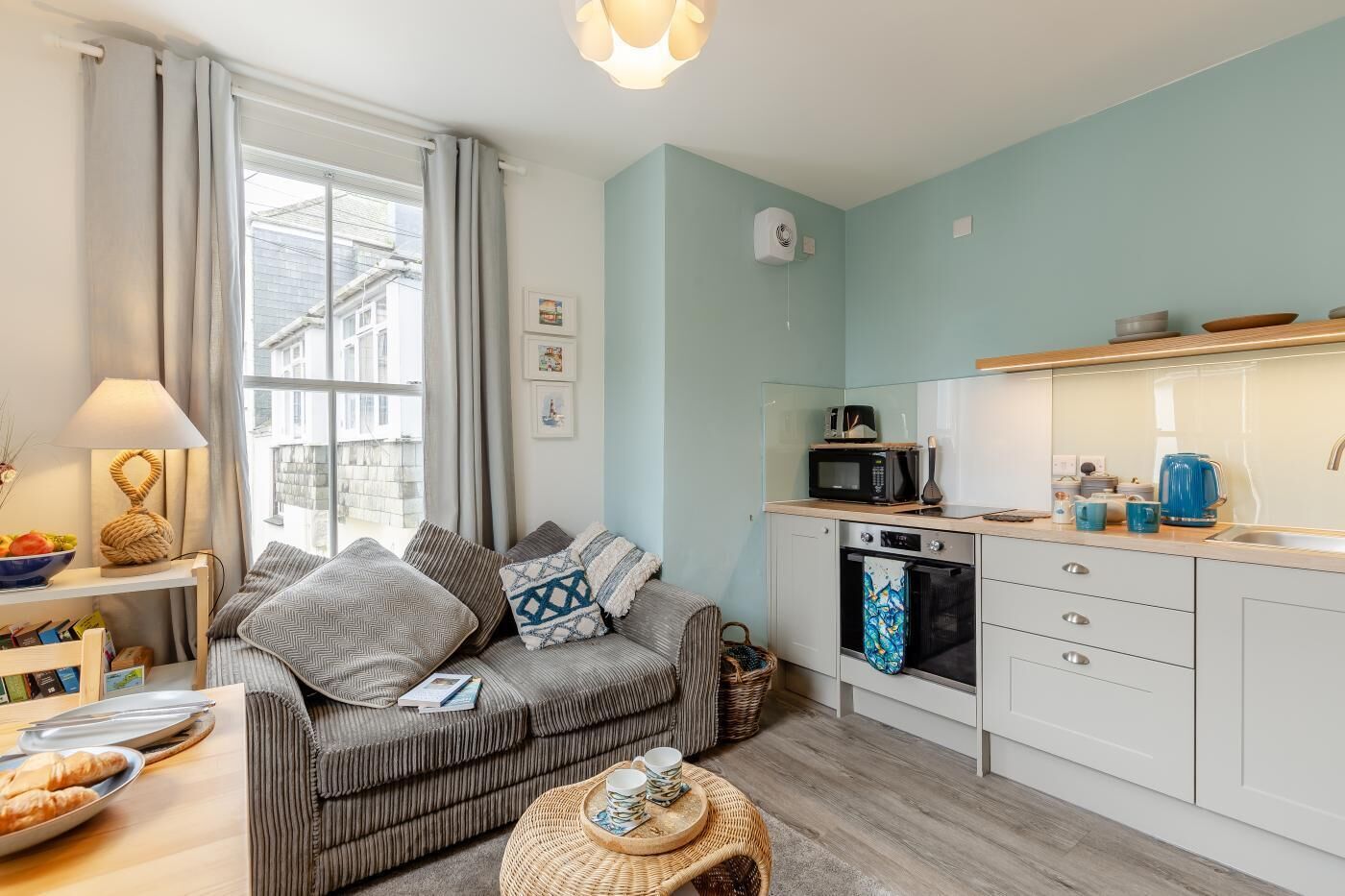 A compact living space with a kitchenette, a sofa with patterned cushions, a dining table with a basket, and a window with curtains.