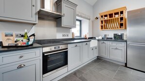 A modern kitchen with grey cabinetry, a built-in oven, a stainless steel refrigerator, and a wooden shelf above the sink. - 1 Bed in Caernarfon (oc-bow270) (Penygroes)