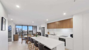 A modern kitchen with a central island, white cabinetry, and a large window providing a view of the city. - The Corso - Central Maroochydore Apartment (Sunshine Coast)