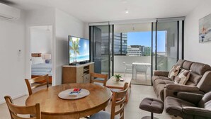 A modern living room with a dining area, a sofa, a TV, and a balcony with a city view. - The Corso - Central Maroochydore Apartment (Sunshine Coast)