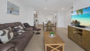 A modern living room with a brown leather sofa, a wooden coffee table, and a TV displaying a beach scene. - The Corso - Central Maroochydore Apartment (Sunshine Coast)