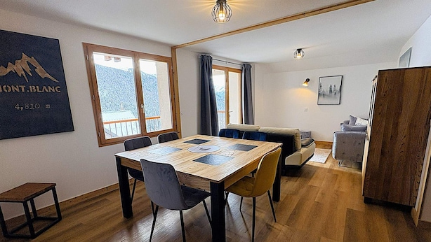 A dining area with a wooden table and chairs, a sofa, and a view of snow-covered mountains through large windows. - Le refuge d'Anny - Three Bedroom Apartment, Sleeps 8 (Vaujany)