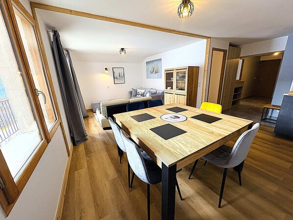 Een eethoek met een houten tafel, zwarte stoelen en een gele stoel. Achterin staat een bank en een kast.