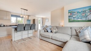 A modern living room with a grey sofa, a dining area with a white table and chairs, and a kitchen in the background. - 3 Bed in Higher Crackington (oc-avond) (Crackington Haven)