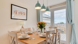 A dining area with a wooden table set for six, white chairs, a vase of flowers, and a framed picture on the wall. - 3 Bed in Higher Crackington (oc-avond) (Crackington Haven)