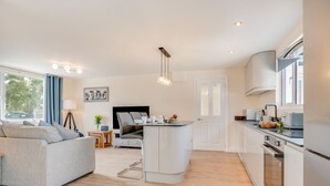 A modern living room with a grey sofa, a dining area with a table and chairs, a kitchen with white cabinets and appliances, and a large window providing natural light. - 3 Bed in Higher Crackington (oc-avond) (Crackington Haven)