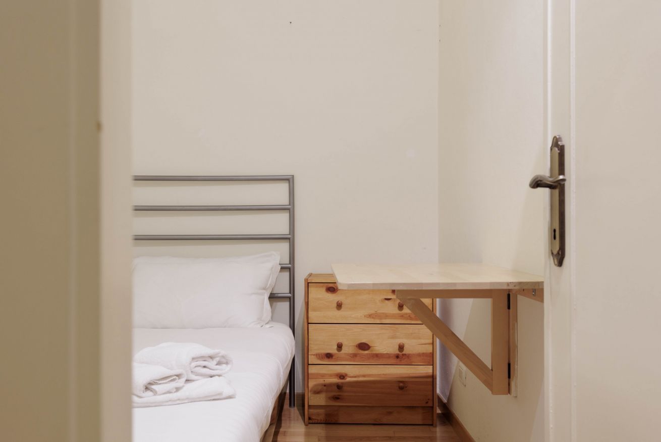 A small, narrow room with a single bed, a wooden chest of drawers, and a small table.