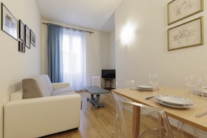 Ein Wohnzimmer mit einer Couch, einem Esstisch für zwei Personen und einem Fernseher im Hintergrund.