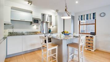 A modern kitchen with white cabinets, a granite countertop, a central island with bar stools, stainless steel appliances, and a hanging light fixture.
