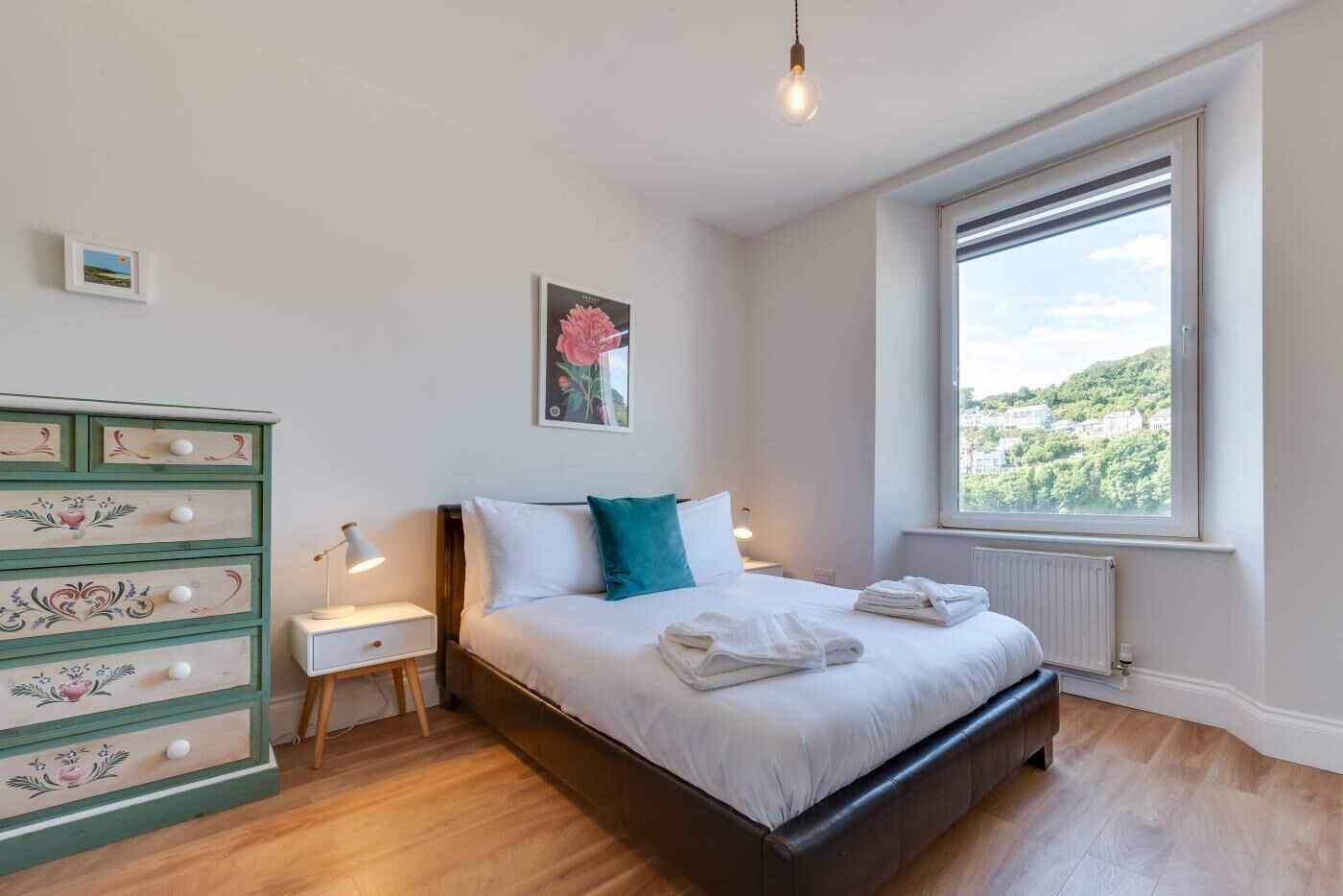 Un dormitorio con cama, cómoda, mesita de noche, ventana con vistas y lámpara colgante.