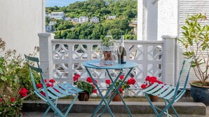 A balcony with two blue chairs, a small table, and potted plants with red flowers. - 1 Bed in Looe (oc-seahol) (Looe)