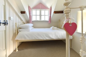 A bedroom with a single bed, white bedding, a window with pink curtains, and a heart-shaped decoration.