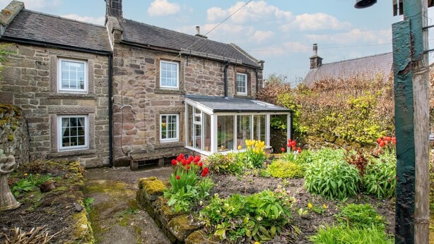 Una casa de piedra con un invernadero, un jardín con diversas plantas y flores, y un cielo despejado.