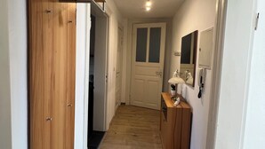 Un pasillo estrecho con puertas de madera a la izquierda, una puerta blanca al final y un pequeño armario de madera con un espejo a la derecha.