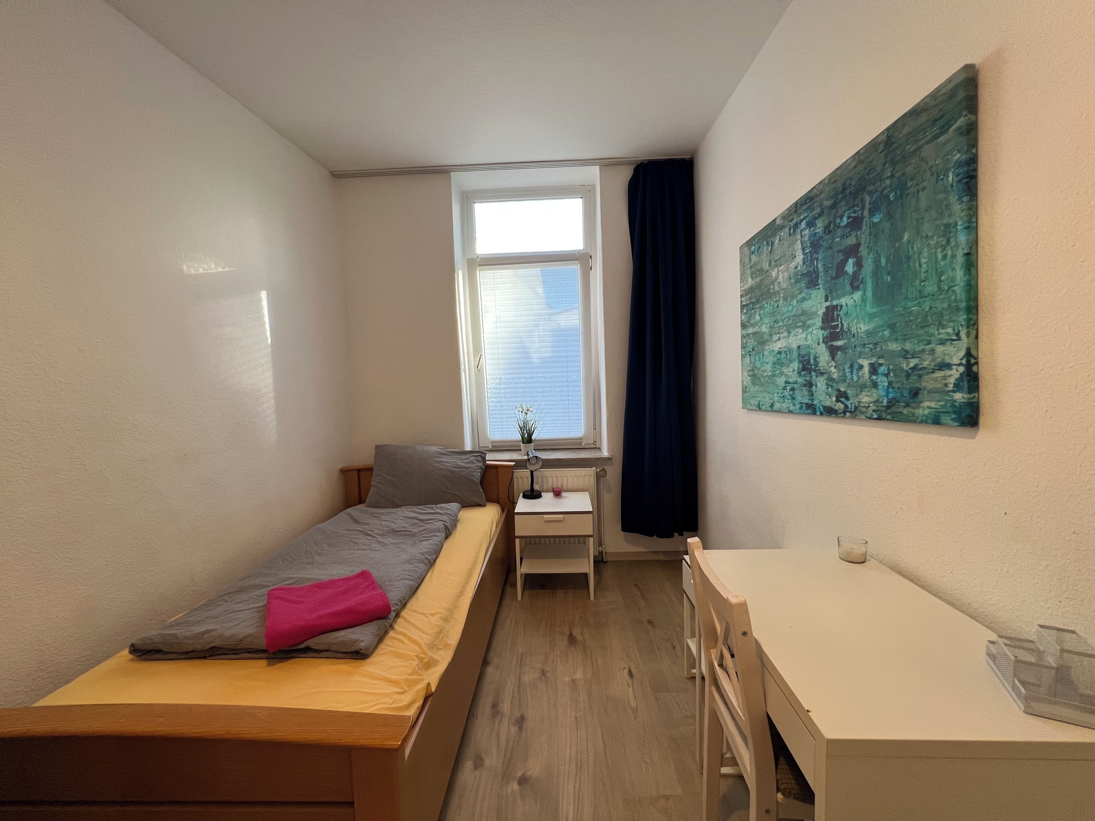 A small, well-lit room with a single bed, a desk, and a radiator. There is a window with blinds and a painting on the wall.
