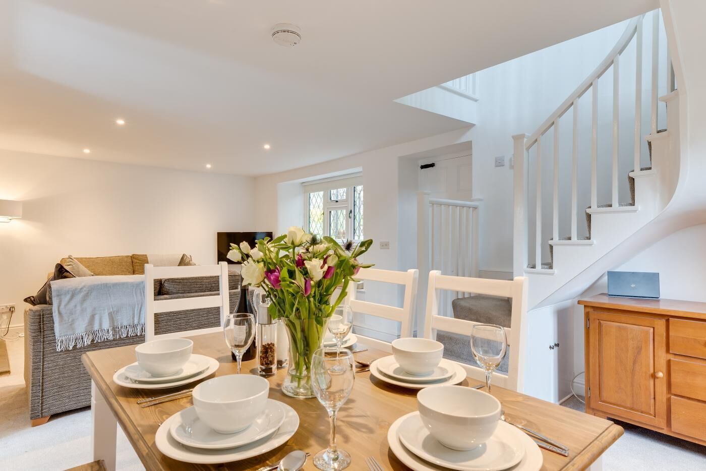 A dining area with a wooden table set for four, white chairs, and a vase with flowers.