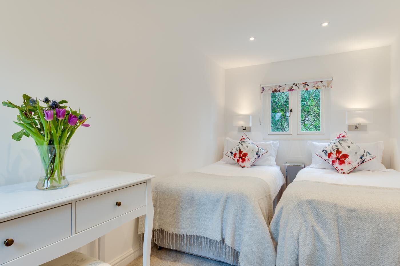 A bedroom with two beds, a nightstand, a dresser, a vase of flowers, and a window with a view of greenery.