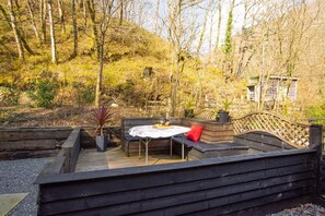 A wooden deck with a built-in seating area and a table, surrounded by a natural landscape with trees and a hill in the background. - 1 Bed in Carmarthen (oc-waw218) (Carmarthen)
