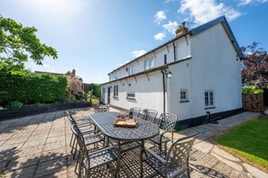 A patio with a round table and chairs, a white building with a chimney, and a hedge in the background. - 4 Bed in Great Plumstead (oc-2295) (Great Plumstead)