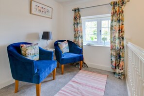 A room with two blue armchairs, a small side table with a lamp, a window with floral curtains, and a framed picture on the wall. - 4 Bed in Great Plumstead (oc-2295) (Great Plumstead)
