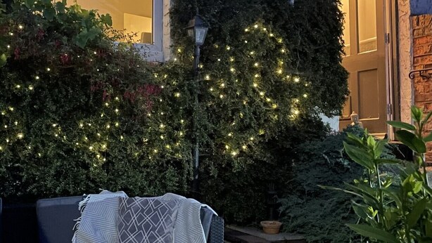 Un espacio al aire libre con mesa, sillas y una manta. Hay plantas y una pared decorada con luces colgantes.