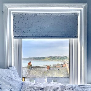 A bedroom with a view of a coastal town through a window with a floral valance. - 3 Bed in Scarborough (oc-s113) (Scarborough)