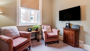 Una sala de estar con dos sillones, una mesita con una planta y un teléfono antiguo, un televisor en la pared y un mueble de madera con una computadora portátil.