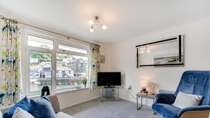 A modern living room with a large window, a flat-screen TV, a blue armchair, and a sofa. - 1 Bed in West Looe (oc-1west) (Looe)