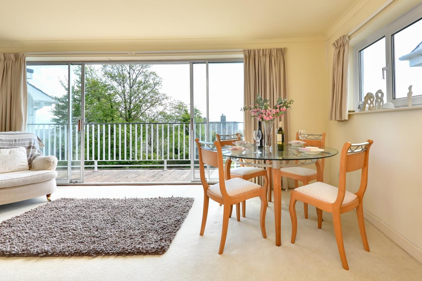 A modern living room with a glass dining table, wooden chairs, a beige sofa, and a large window with a balcony view.