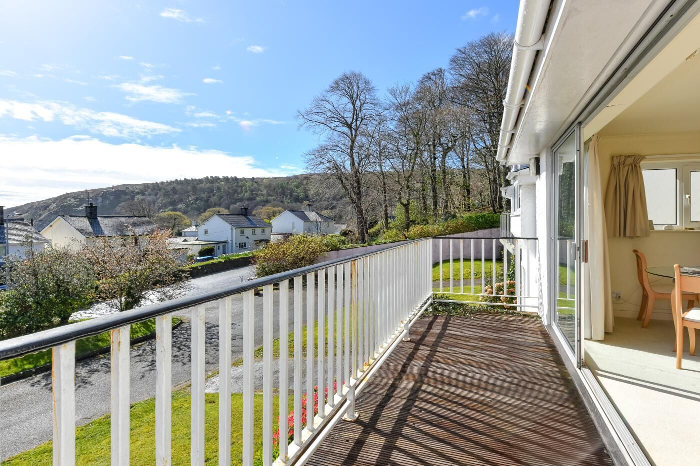 A balcony with a view of houses and trees, a wooden deck, and a glass door leading to a room with a table and chairs.