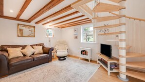 Una sala de estar con un sofá de cuero marrón, un sillón blanco, una escalera espiral de madera, un mueble de TV blanco y un pequeño calefactor blanco.