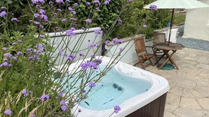 Un jacuzzi rodeado de exuberante follaje y flores púrpuras, con sillas de madera y una mesa cerca.