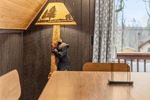 A dining area with a table, chairs, a lamp with a forest design, and a bear figurine leaning on a wooden post.