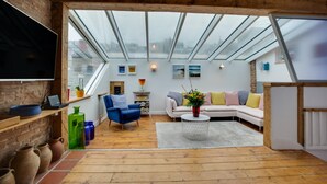 A modern loft with a large skylight, wooden flooring, a white sofa, a blue armchair, and a flat-screen TV. - 5 Bed in St. Ives (oc-baktre) (St Ives)