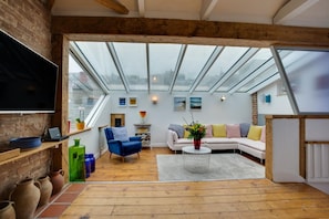 Ein modernes Loft mit großem Dachfenster, Holzboden, einem weißen Sofa, einem blauen Sessel und einem Flachbildfernseher.
