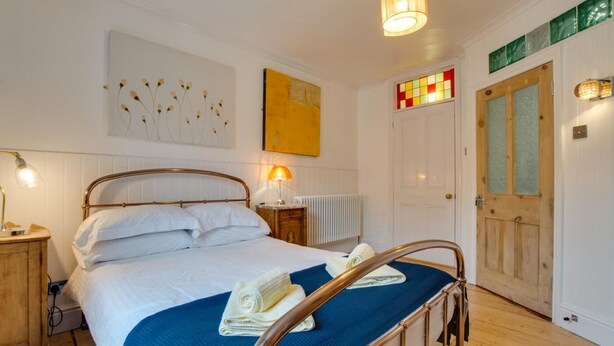 A bedroom with a metal-framed bed, white bedding, a blue throw blanket, a rolled towel, a bedside lamp, a radiator, a wooden nightstand, a wall with a floral painting, a door with a stained glass window, and a green sign above the door. - 5 Bed in St. Ives (oc-baktre) (St Ives)