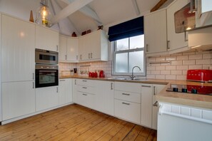 A kitchen with white cabinets, a built-in oven, a window with a blue valance, and a red toaster. - 5 Bed in St. Ives (oc-baktre) (St Ives)