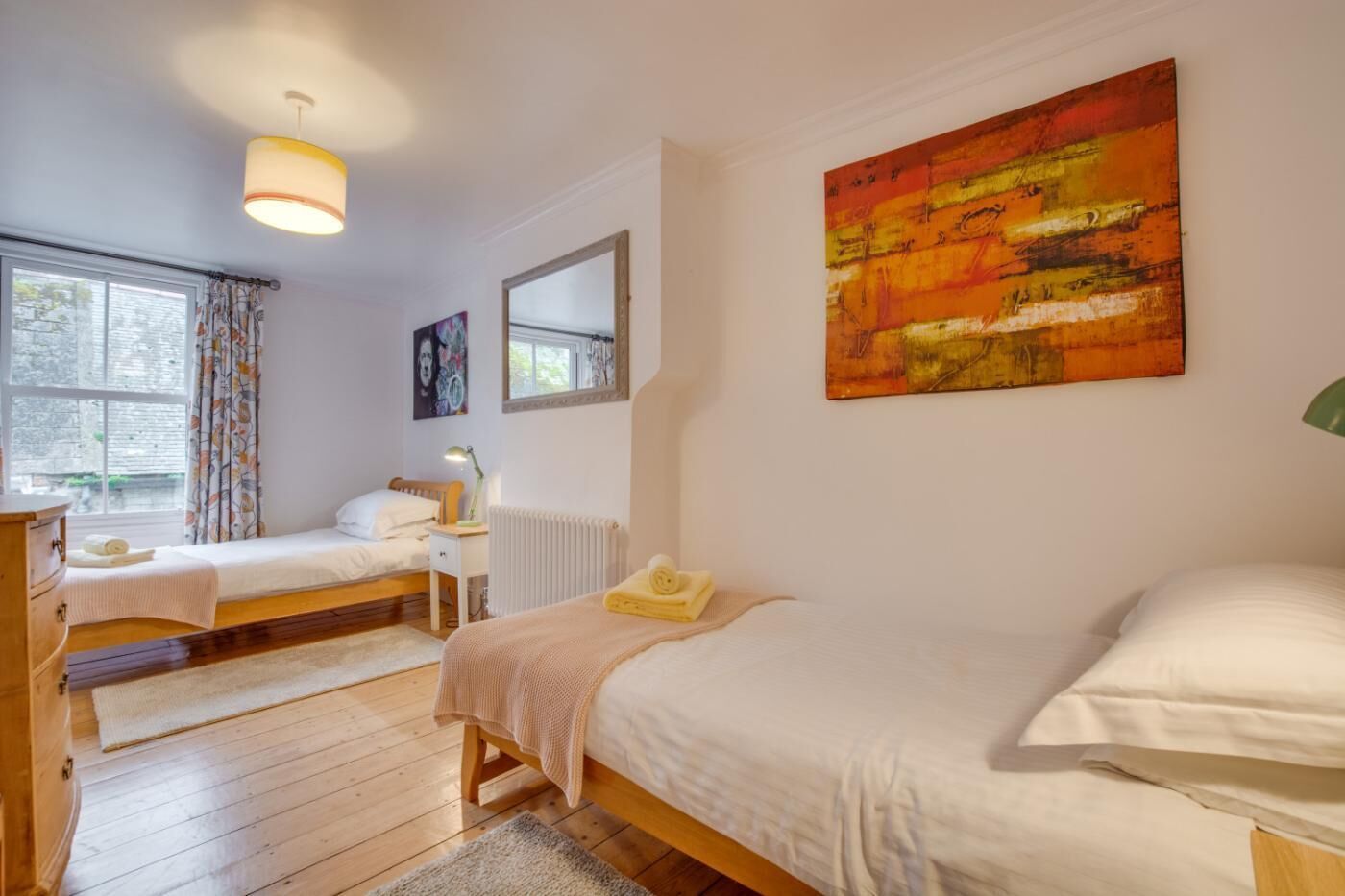 A bedroom with two single beds, a window with curtains, a radiator, and a painting on the wall.