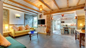 A cozy living area with a sofa, a coffee table, and a TV. There's a stone wall and a kitchen in the background. - 5 Bed in St. Ives (oc-baktre) (St Ives)