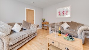 A living room with two grey sofas, a wooden coffee table with a bottle and glasses, and a framed floral artwork on the wall. - 2 Bed in Fowey (oc-jackda) (Fowey)