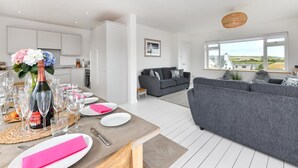 A modern living room with a grey sofa, a dining table set for a meal, and a kitchen in the background. - 4 Bed in Abersoch (oc-21tan) (Abersoch)