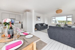 A modern living room with a grey sofa, a dining table set for a meal, and a kitchen in the background. - 4 Bed in Abersoch (oc-21tan) (Abersoch)