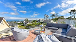 A rooftop terrace with a grey sofa, a white coffee table with a floral centerpiece, and a grey bean bag. - 4 Bed in Abersoch (oc-21tan) (Abersoch)