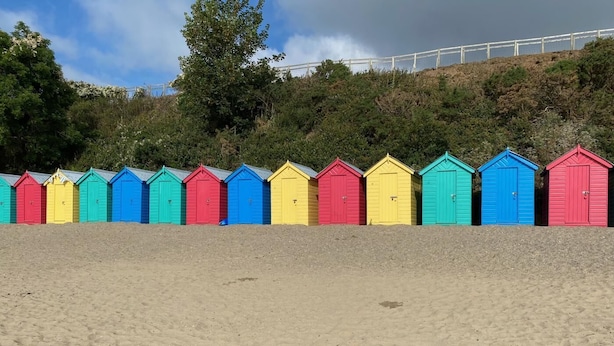 Eine Reihe bunter Strandhäuser am sandigen Strand, im Hintergrund ein Hügel und Bäume.