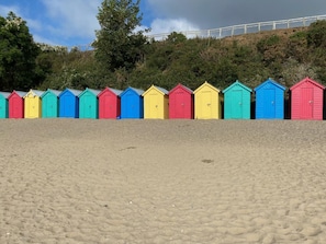 En rad färgglada strandhytter längs en sandstrand, med en kulle och träd i bakgrunden.