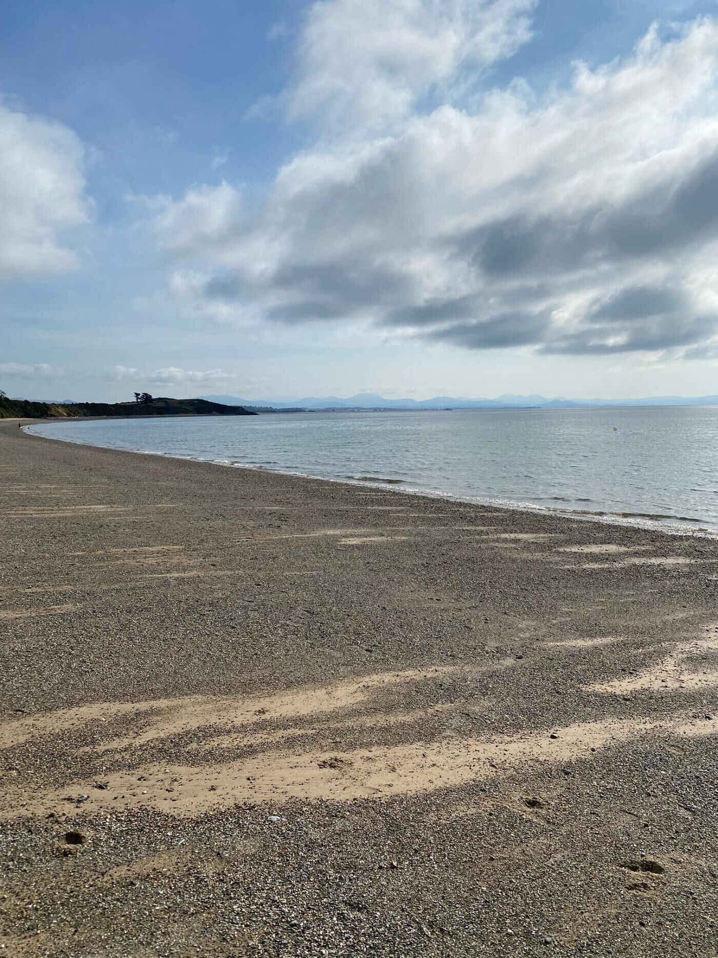 Una playa con piedras y arena, cielo despejado y montañas a lo lejos.